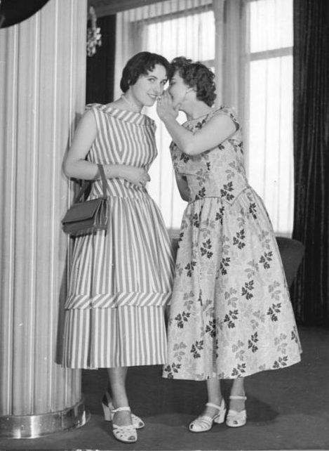 picture of two women in party dresses from 1956, one whispering in the ear of the other